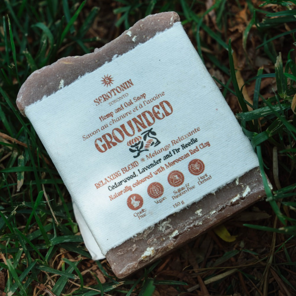 A bar of Serotonin 'Grounded' handmade soap resting on a bed of green grass and brown leaves. The soap has a rustic, rough texture with a white paper label wrapped around it, featuring brown text and illustrations.