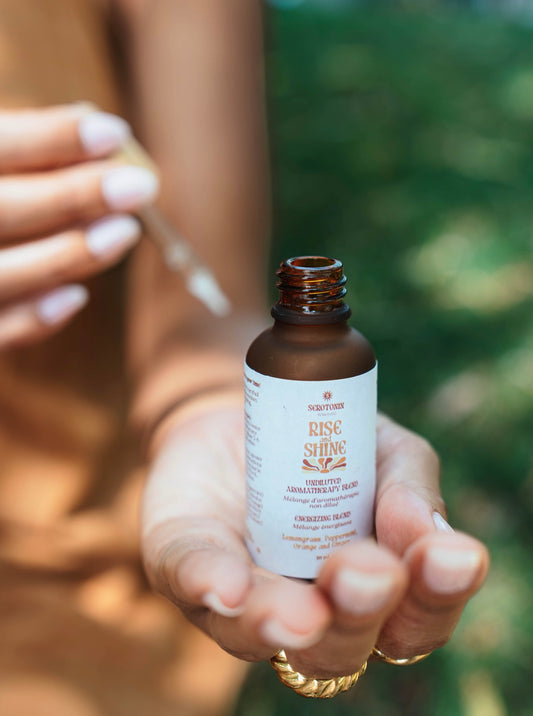 A person holds an amber glass bottle labeled Rise Shine Aromatherapy blend in their hand, with a glass dropper above it, against a blurred green outdoor background.