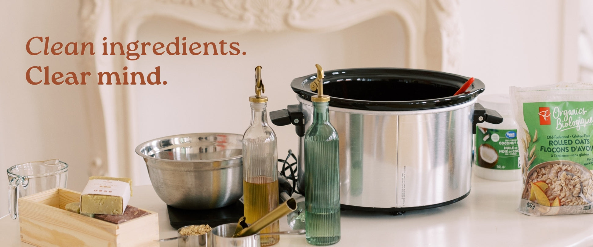 A stainless steel slow cooker sits on a white table alongside various kitchen ingredients and utensils, with the text 'Clean ingredients. Clear mind.
