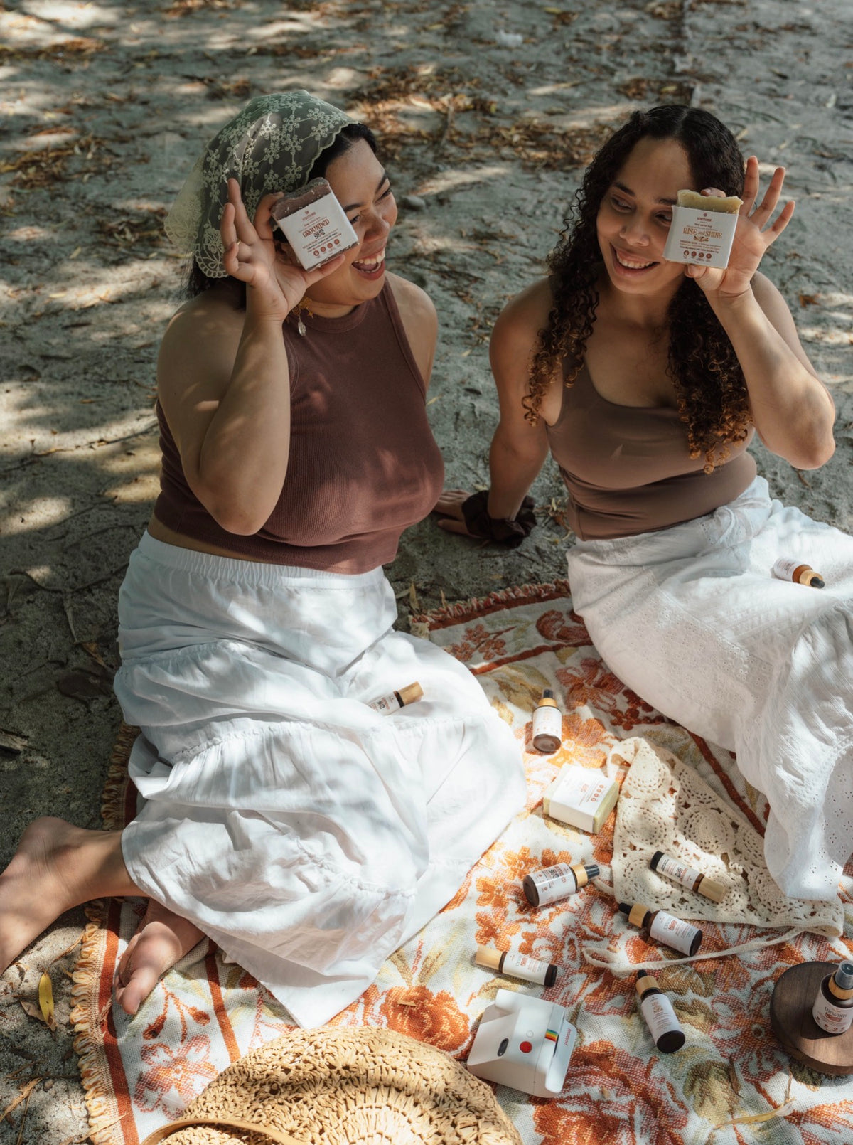 Two women sitting on a blanket outdoors, holding up small white product boxes. The scene includes several small cylindrical bottles and containers scattered on the blanket, with a straw hat in the foreground and dappled sunlight on the ground.