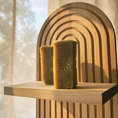 Two rectangular handmade soap bars with embossed sun ray designs, displayed on a wooden shelf in front of a wooden arch backdrop.