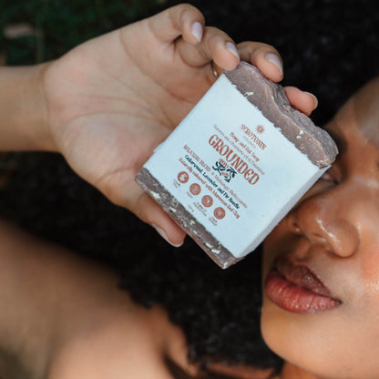 A close-up lifestyle shot of a person holding a bar of Soap of the Gods 'Grounded' soap bar. The hand holds the bar, which has a white paper label with brown text, against a dark, blurred background of the person's face and hair.