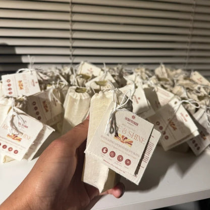 A hand holds a small cream-colored fabric pouch with a paper tag labeled 'SEROTONIN' and 'RISE & SHINE', with many similar pouches arranged on a shelf behind it in front of window blinds.