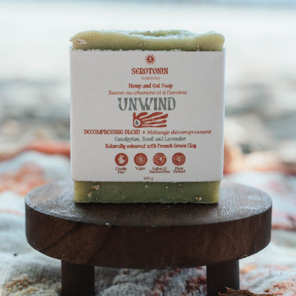 A bar of Serotonin Unwind Hemp and Oat Soap sits on a small round wooden stool. The soap bar is wrapped in a white paper label with red and green text, and the top and bottom pieces are a natural light green.