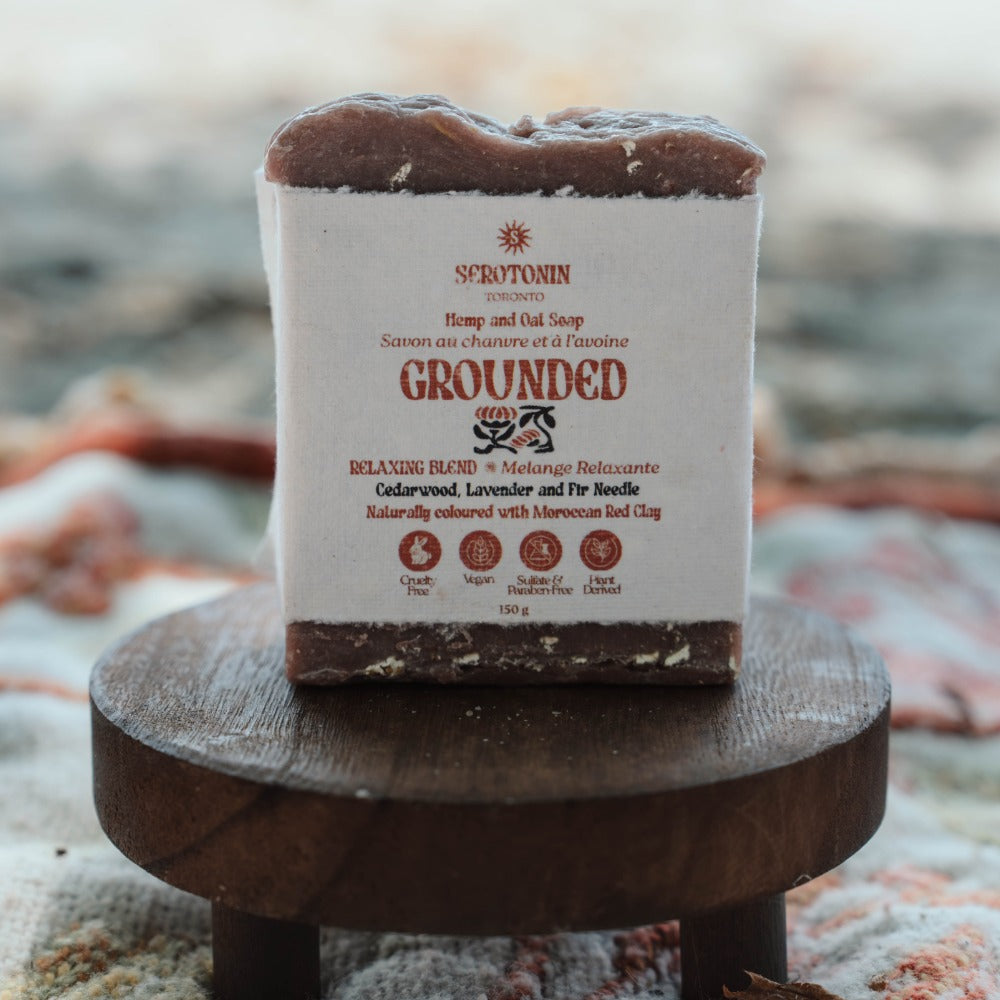 A rectangular bar of soap labeled 'SEROTONIN GROUNDED' sits on a small round wooden stand. The soap has a white paper label with brown and black text, and the top and bottom are a dark brown natural color.