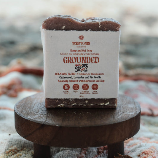 A rectangular bar of soap labeled 'SEROTONIN GROUNDED' sits on a small round wooden stand. The soap has a white paper label with brown and black text, and the top and bottom are a dark brown natural color.