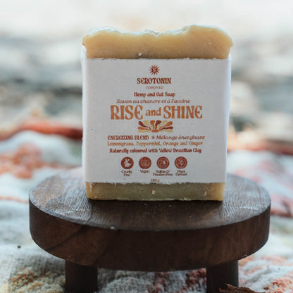 A rectangular bar of handmade soap labeled 'Rise and Shine' by Serotonin Toronto, resting on a small round wooden stand. The soap has a white paper label with brown text and graphics, and a natural beige-tan color visible at the top and bottom edges.