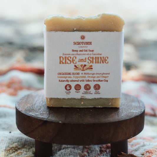 A rectangular bar of handmade soap labeled 'Rise and Shine' by Serotonin Toronto, resting on a small round wooden stand. The soap has a white paper label with brown text and graphics, and a natural beige-tan color visible at the top and bottom edges.