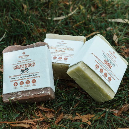 Three bars of soap from Serotonin Toronto are arranged on a grassy ground with dry leaves. The soaps are wrapped in white paper labels, showing their natural brown and light green solid bars.
