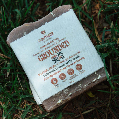 A bar of Serotonin 'Grounded' handmade soap resting on a bed of green grass and brown leaves. The soap has a rustic, rough texture with a white paper label wrapped around it, featuring brown text and illustrations.